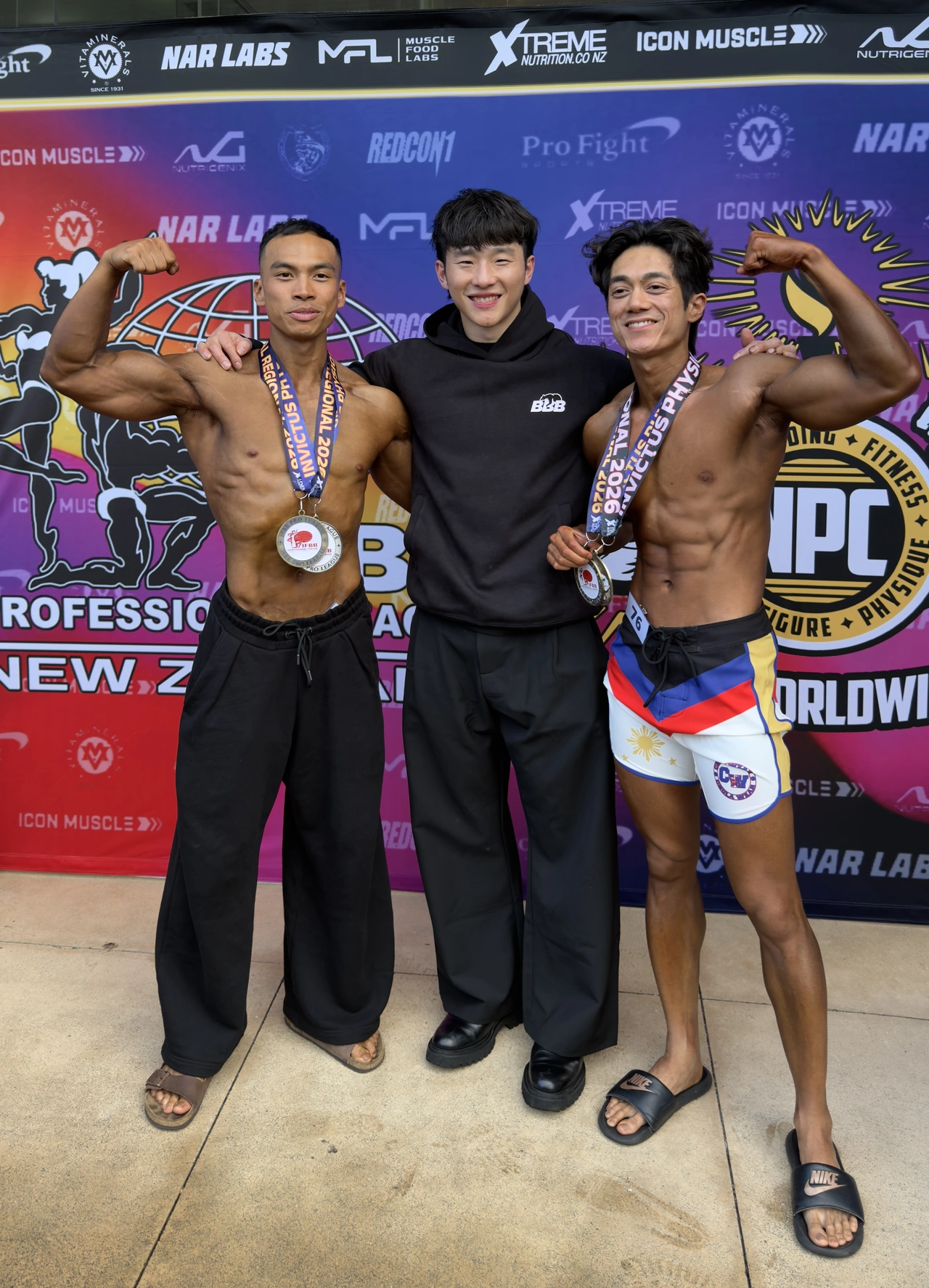 Boxi standing with two competitors wearing medals in front of a colourful event backdrop.
