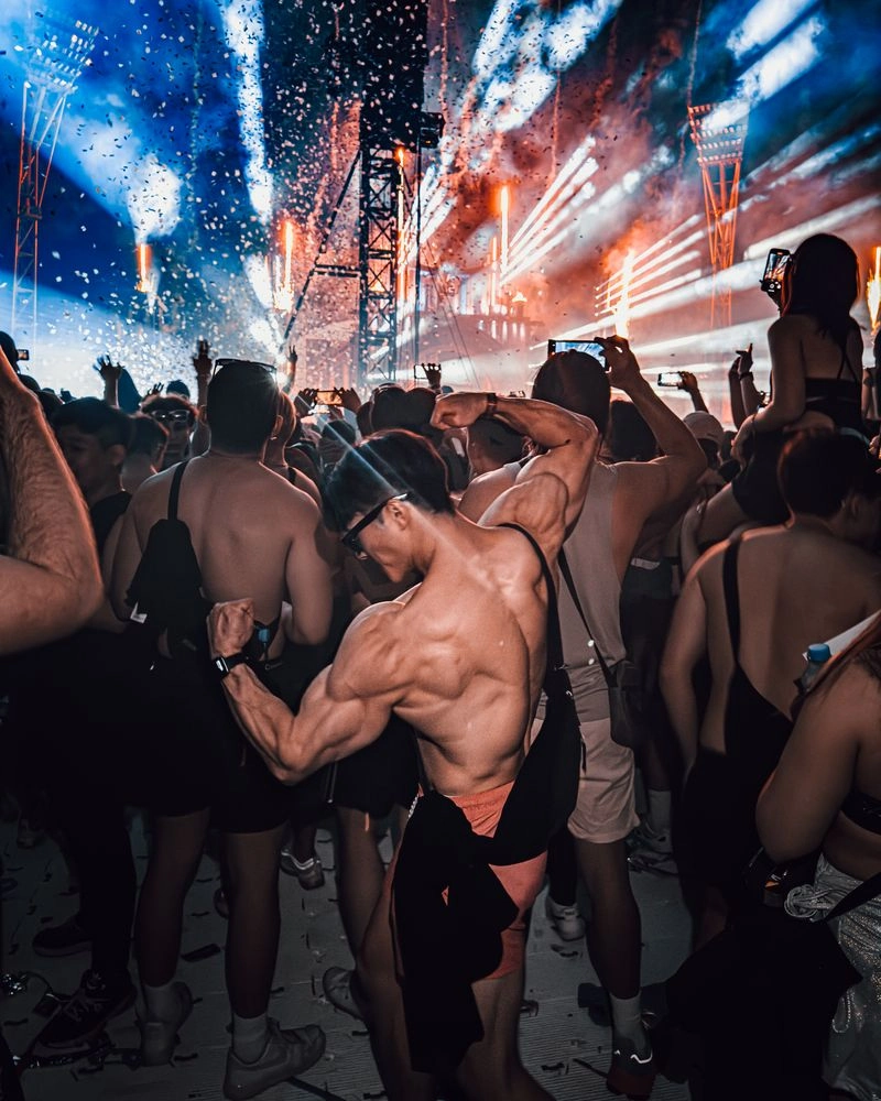 Boxi striking a back double-biceps pose at a rave under stage lights and confetti.